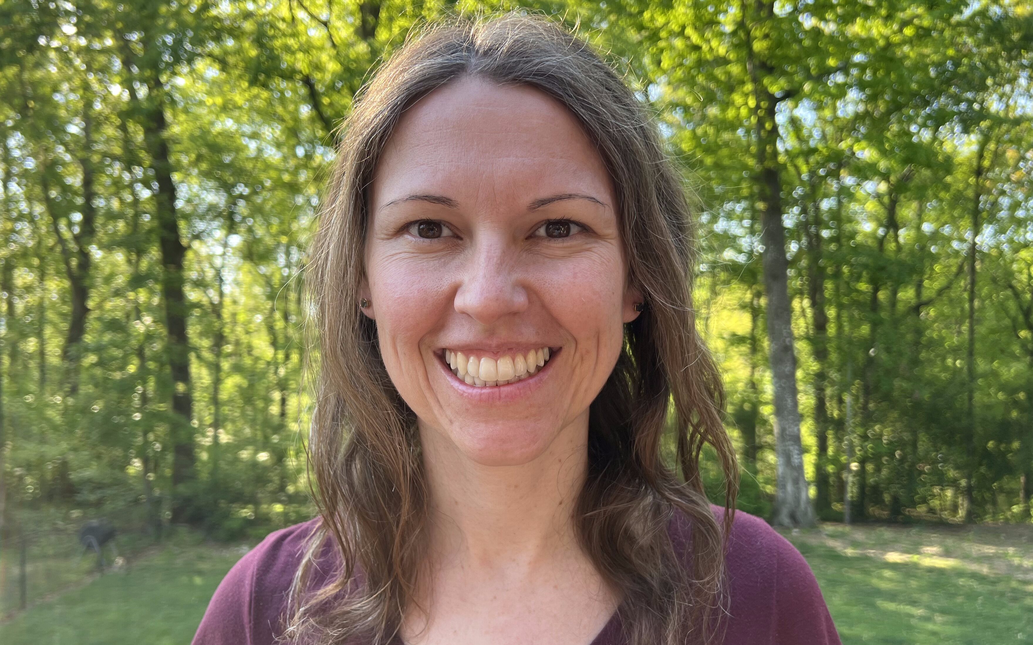 Elizabeth Beu Bourgeois wearing a grey shirt and glasses in front of a vine covered wooden fence